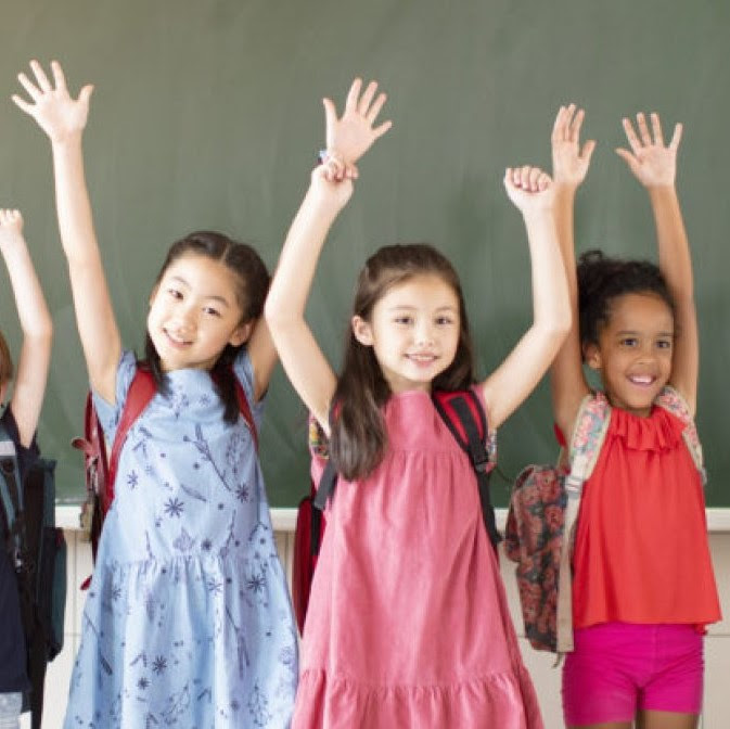 three young kids wearing backpacks raise their arms in the air in front of a blackboard