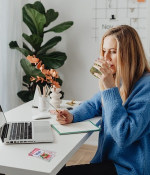 Woman in Blue Sweater at Desk with Laptop Drink from Clear Glass with Liquid Packets of Rebellious Infusions Strawberry Kiwi Nearby