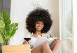A smiling Black woman with a large Afro sits near a bright window, reading a book and drinking a gla...