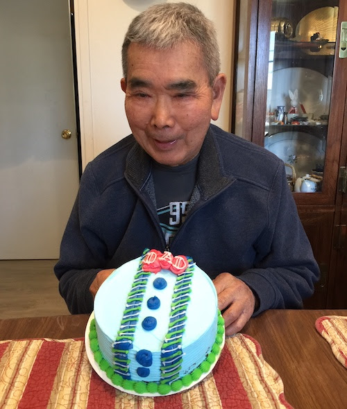 Image of older man with short grey hair and a dark blue jacket in front of a cake that says Dad