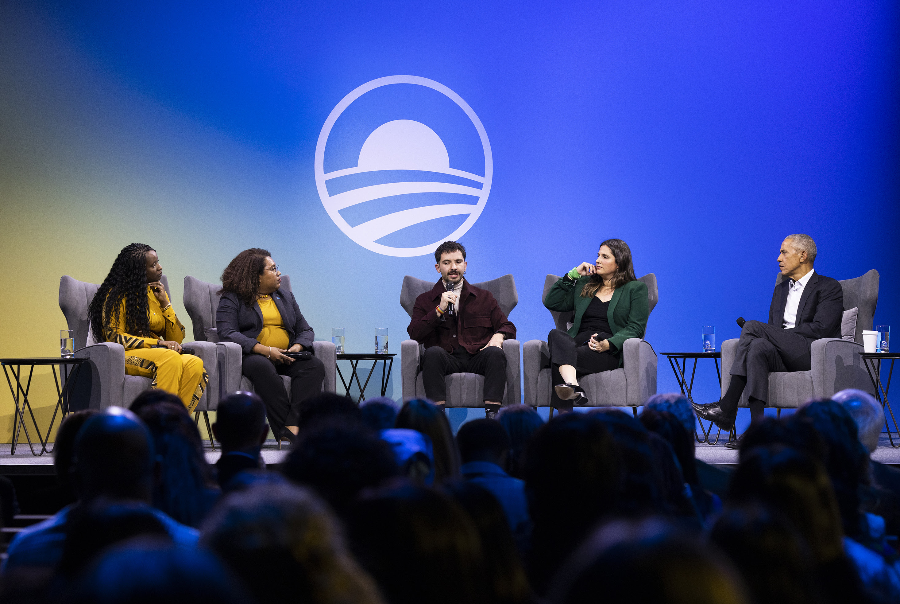 Nick Antipov sits on stage and speaks into a microphone on a panel alongside Doussouba Konaté, Landisang Kotaro, Natalia Herbst, and President Obama. All the speakers are a range of light and deep skin tones. In the background is the Obama Foundation logo, an abstract rising sun in the shape of the letter O. 