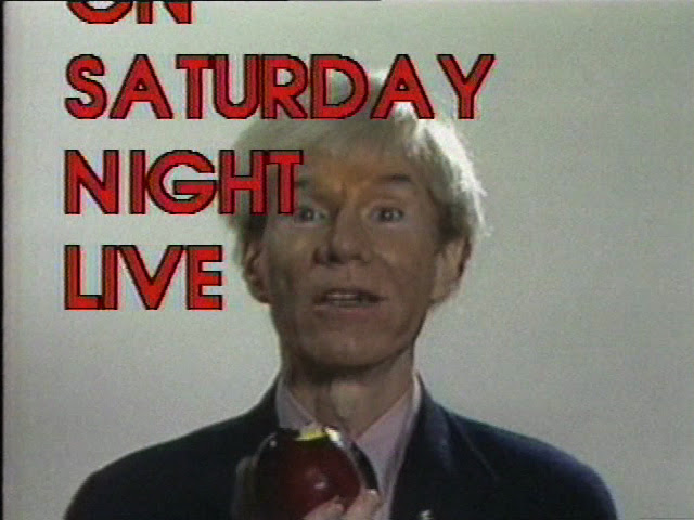 A video still of a person holding an apple and looking towards the camera. A "Saturday Night Life" graphic in red, capital letters appears in the foreground.