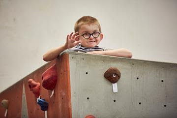 A boy climbing an indoor wall and waving at the camera