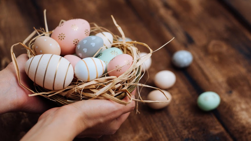 A pair of hands holds a nest of eggs. They have been dyed pastel colors like Easter eggs, and have interesting patterns in gold foil.