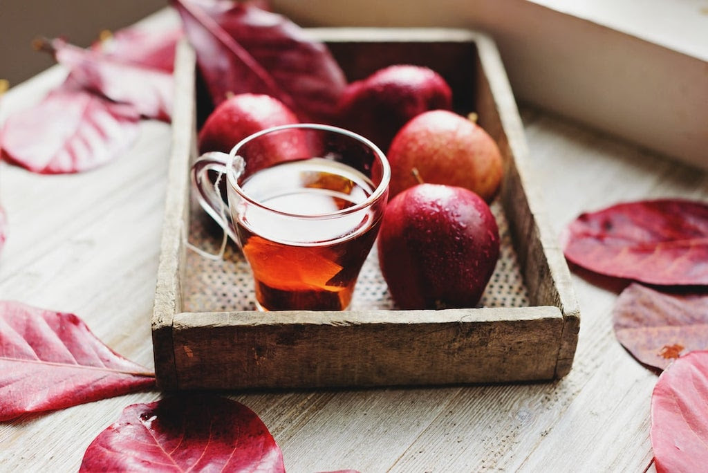 A mug of apple cider and some glistening pinkish red apples on a rustic wooden tray resting on a wood table surrounded by crimson leaves.