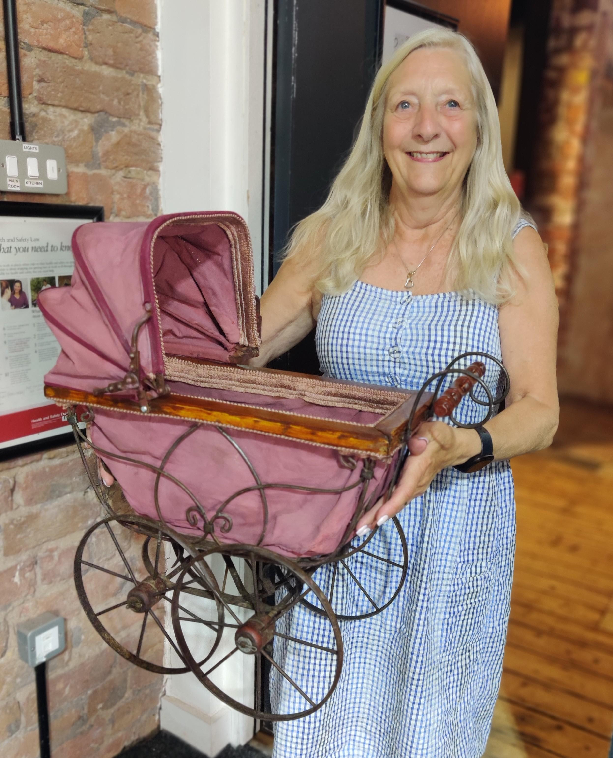 A light haired woman wearing a sundress and holding up an sntique-looking toy pram.