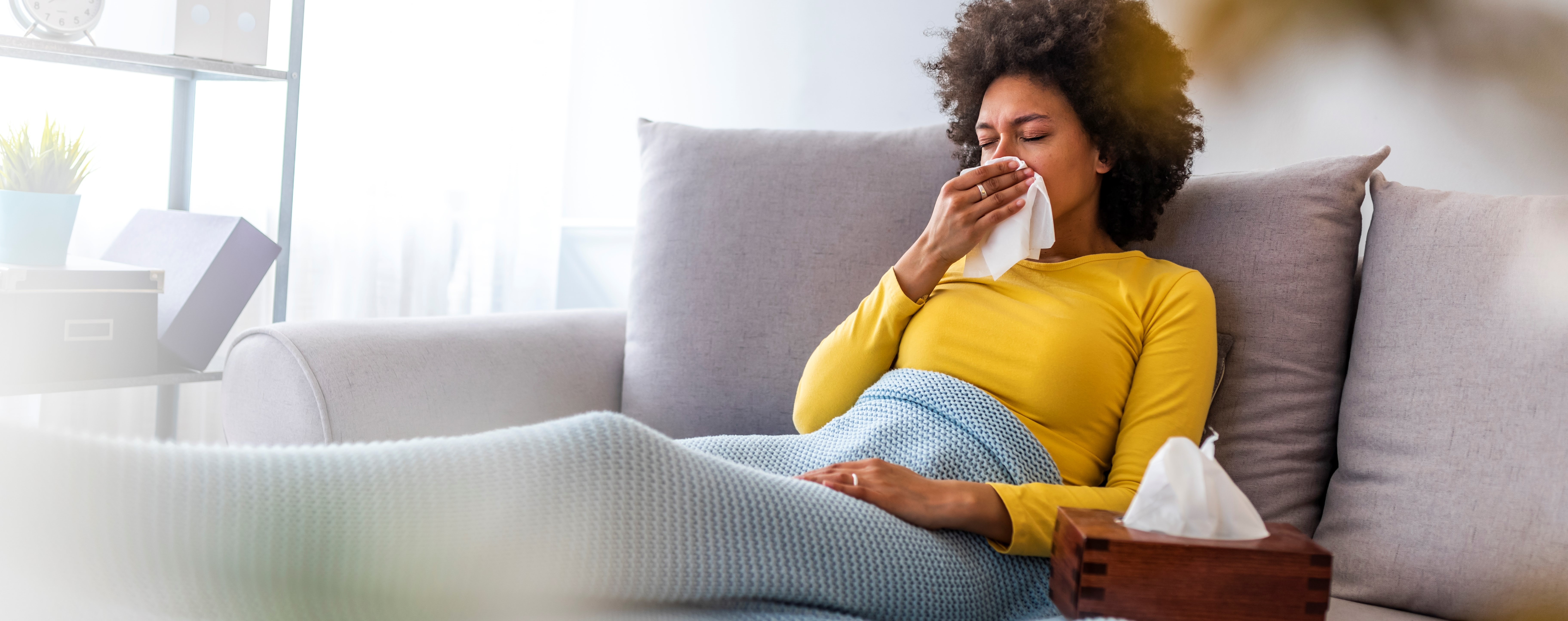 A woman sits under a blanket on her couch blowing her nose. She has a box of tissues sitting next to her.