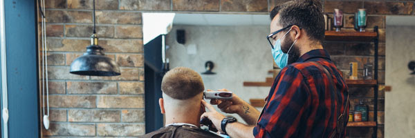 a barber cuts a clients hair while wearing a face mask