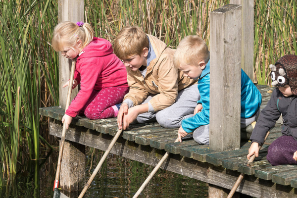 Four young children kneeling on the side of a bridge pushing rods into a lake with smiles on their faces having fun.