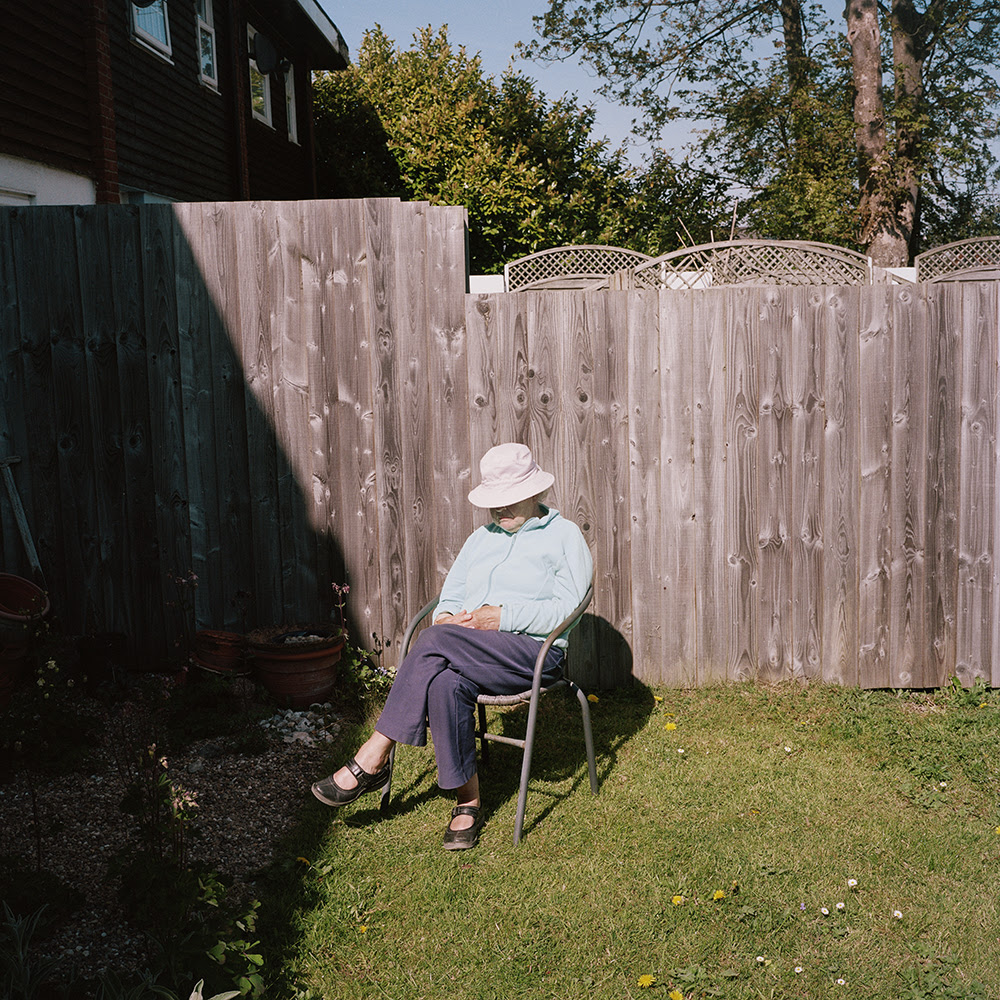[IMAGE] The second image shows an older woman sat in a garden chair in a sunny garden. She is wearing a light blue shirt, navy trousers and a white hat.