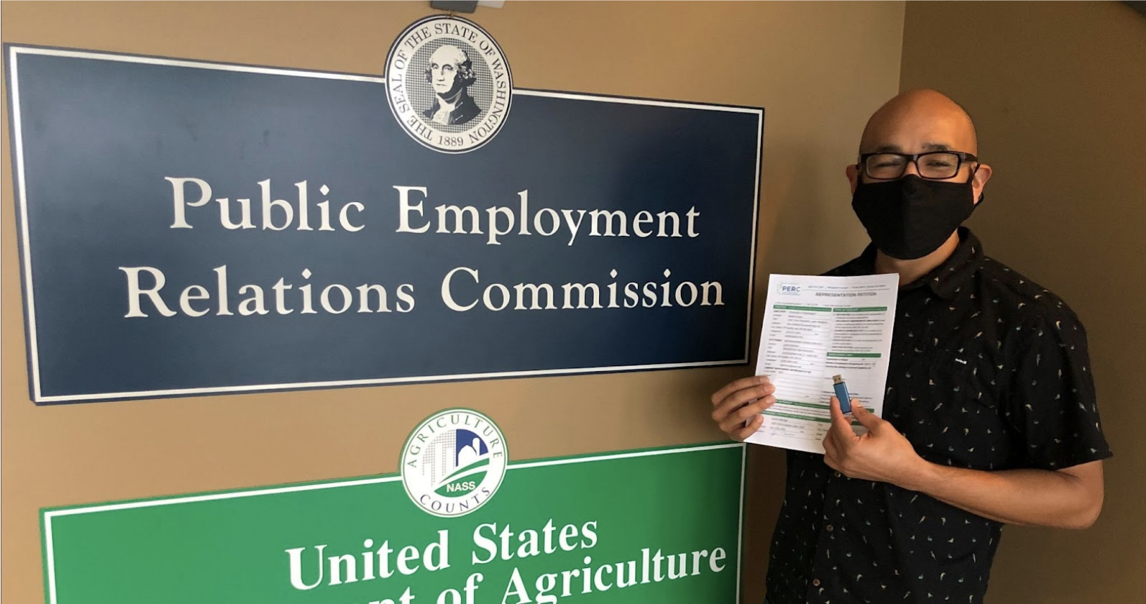 Smiling person with a face mask holding up authorization cards in front of a sign that says "Public Employment Relations Commission"