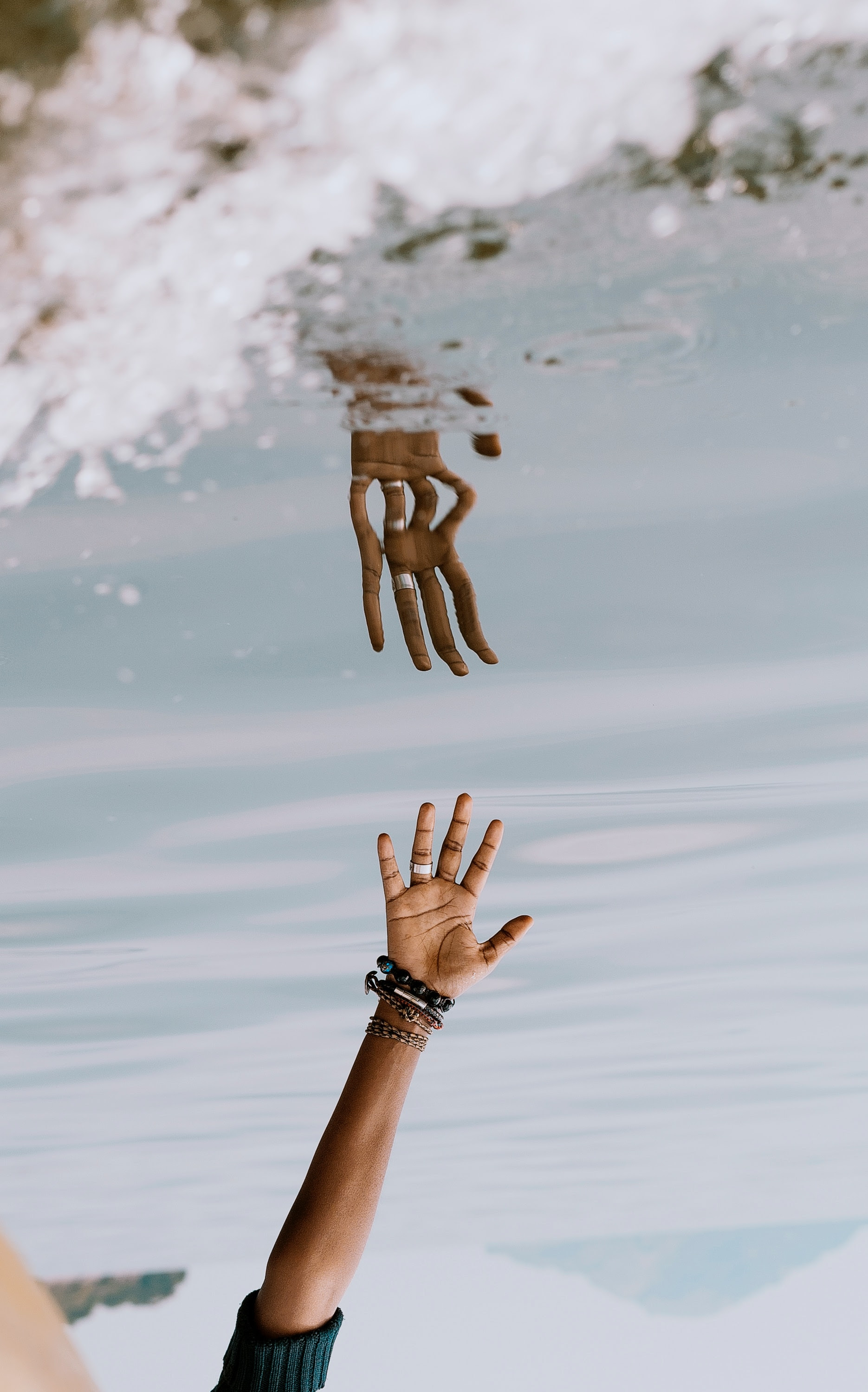 Hand reaching toward the water and being reflected back as it almost reaches its reflection. Photo by Serrah Gallos via Unsplash.