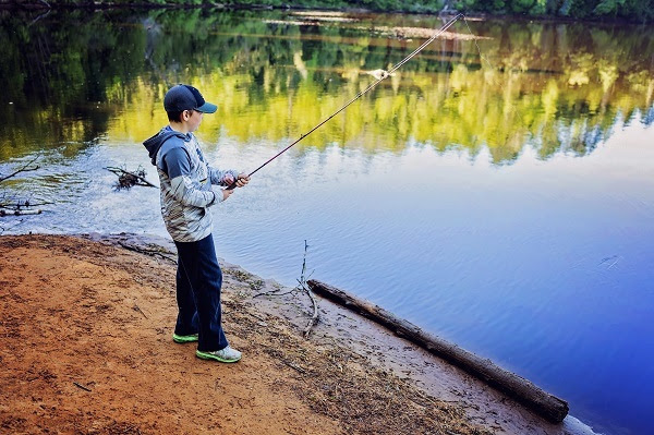 a young boy in jeans, jacket and blue baseball cap stands on shore, holding a fishing rod with line cast close by in calm water