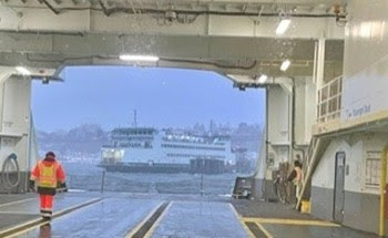 View of car deck of ferry from behind windshield of vehicle with crewmember walking through the tunnel and another ferry in the background